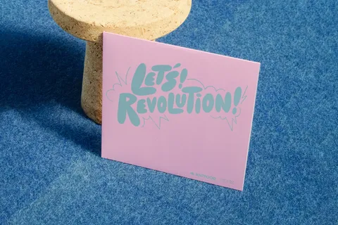 The pink sleeve propped against a cork stool on blue carpet, its logo in pale teal
