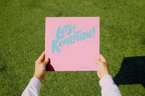 Two hands holding up the Let's! Revolution! record over grass, the pink sleeve showing the game's logo